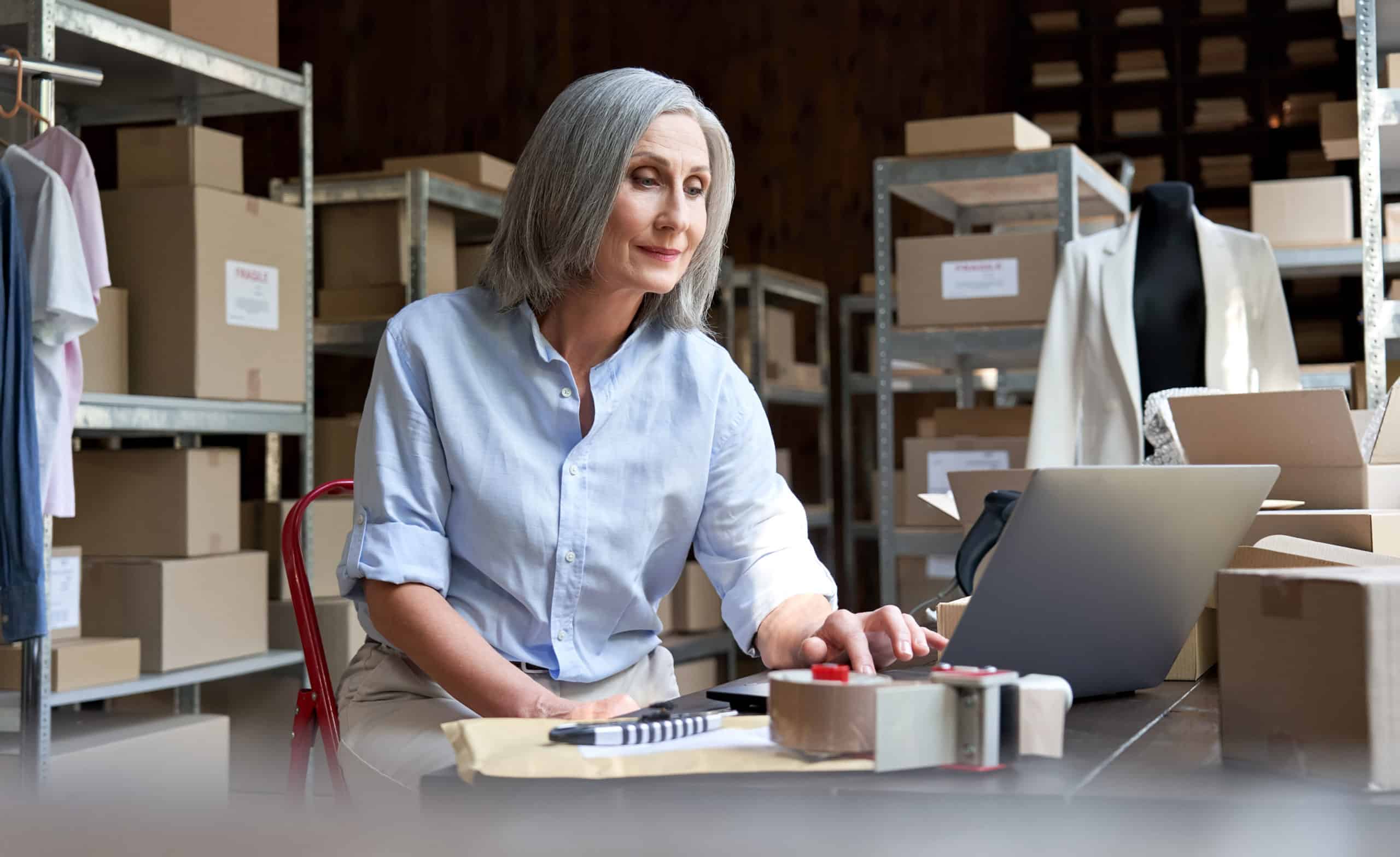 Mature female seller using computer checking ecommerce clothing orders. Maturefemalefashionsellerusingcomputercheckingecommerceclothingstoreorders.Oldermiddleagedbusinesswomanentrepreneurworkingonlaptoppreparingonlineshippingdeliveryparcelsboxes. scaled