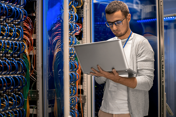 Portrait of modern young man holding laptop standing in server room working with supercomputer in blue light